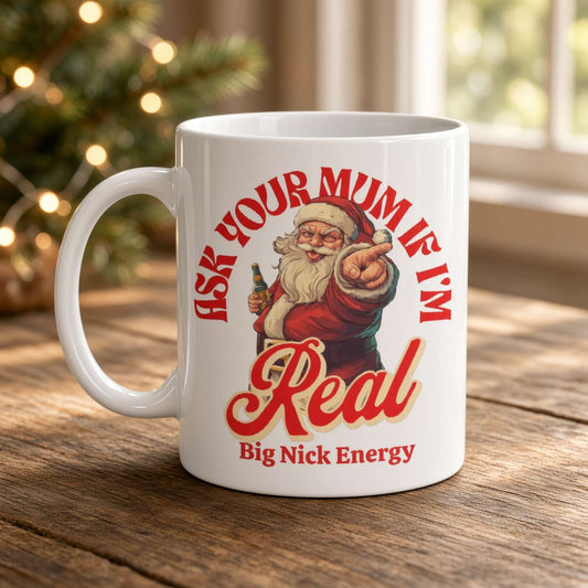 A white mug on a wooden table shows a winking Santa and the playful slogan “Ask your mum if I’m real—Big Nick Energy,” with festive lights glowing behind it.