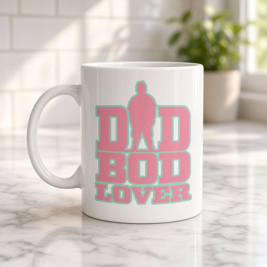 A white ceramic mug on a marble counter reads “DAD BOD LOVER” in bold pink and teal lettering, with a dad silhouette; warm, cheerful kitchen setting.