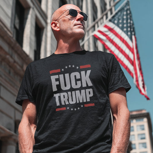 Man wearing a black t-shirt with 'FUCK TRUMP' text in an urban setting with an American flag.