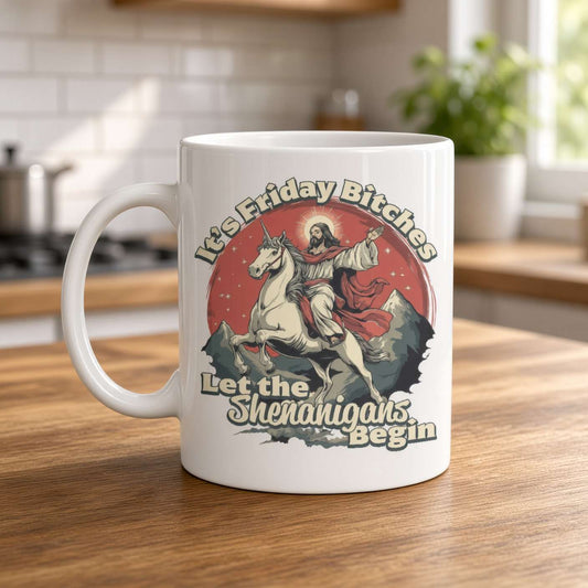 A white ceramic coffee mug with a humorous graphic print, sitting on a wooden table in a blurred kitchen setting. The graphic features a cartoon illustration of Jesus riding a unicorn in front of a red sun, with the text 'It's Friday Bitches Let the Shenangars Begin'.