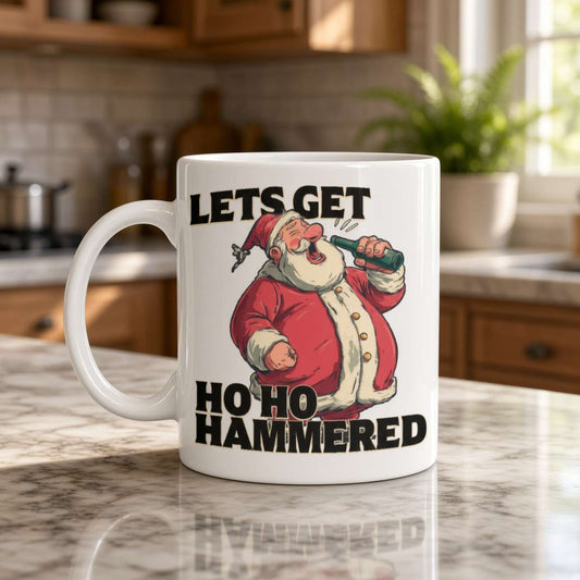 A white mug on a sunlit kitchen counter shows a cheerful, rosy-cheeked Santa holding a drink beneath the bold joke, “Let’s get ho ho hammered.”