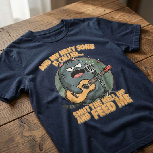 Navy blue t-shirt laid flat on a wooden table showing the full angry cat musician design and the witty “And My Next Song Is Called… Shut The Hell Up And Feed Me” text