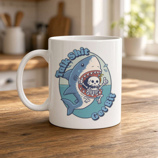 A white ceramic mug on a wooden kitchen counter features a cartoon shark swallowing a skeleton and playful blue lettering, giving a quirky, humorous feel.