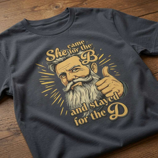 Ink grey She Came for the B and Stayed for the D t-shirt laid flat on a wooden surface showing the full bearded man graphic, funny graphic design full of pure attitude