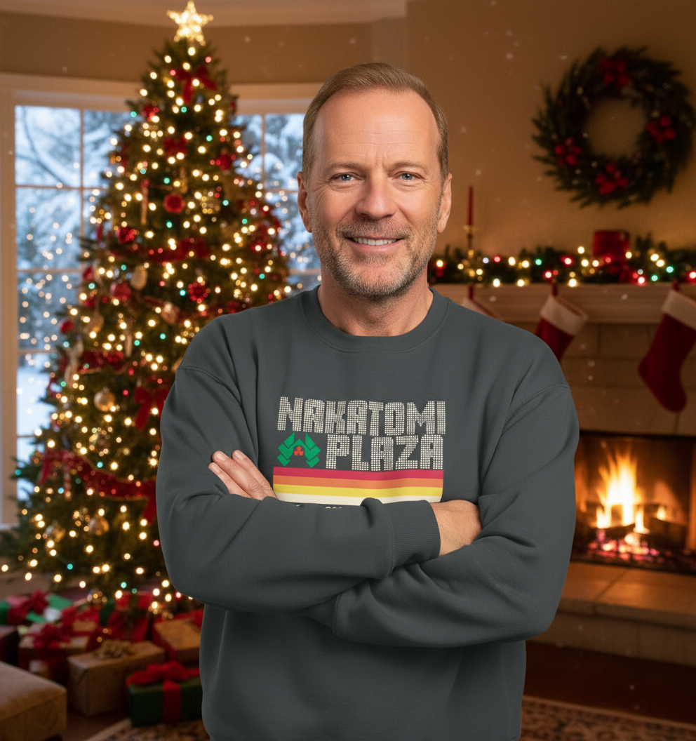 Man wearing a sweater with 'Nakatomi Plaza' design in a festive living room with Christmas tree and fireplace.