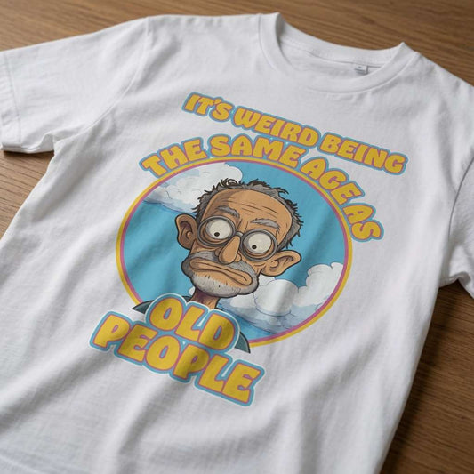 White It’s Weird Being The Same Age As Old People t-shirt laid flat on a wooden surface showing the full cartoon old man graphic, hilarious mens dad gift graphic design full of pure attitude