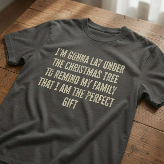 Khaki green t-shirt laid flat on a wooden table with the cream slogan “I’m Gonna Lay Under The Christmas Tree To Remind My Family That I Am The Perfect Gift”, fun and witty mens Christmas pub day design
