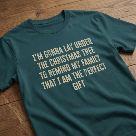 Teal green t-shirt laid flat on a wooden surface with the cream slogan “I’m Gonna Lay Under The Christmas Tree To Remind My Family That I Am The Perfect Gift”, funny and witty ladies xmas banter design