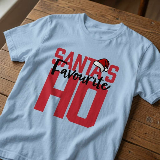 Light blue Santa’s Favourite HO t-shirt laid flat on a wooden surface with bold red and black text, fun Christmas banter design