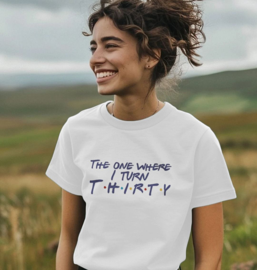 Smiling woman in a white t-shirt with "The One Where I Turn Thirty" in playful text. She stands in a field under a cloudy sky, conveying joy and excitement.