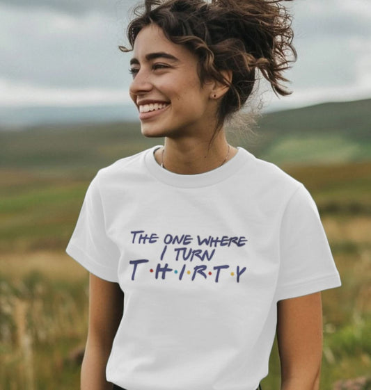 Smiling woman in a white t-shirt with "The One Where I Turn Thirty" in playful text. She stands in a field under a cloudy sky, conveying joy and excitement.