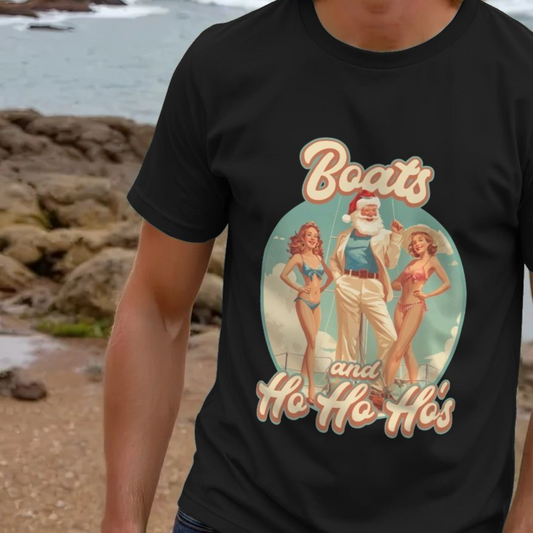 Man wearing a black funny Christmas t shirt with a retro-style graphic of Santa between two women in bikinis, reading "Boats and Ho Ho Ho's." Beach background visible.