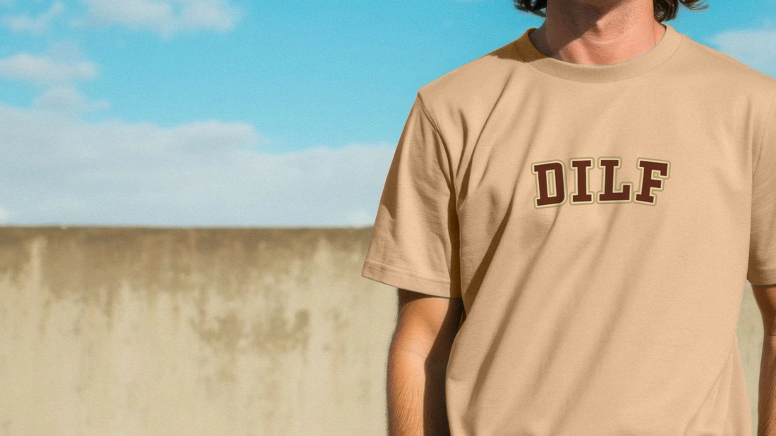 A person wearing a tan funny Dad T-shirt with "DILF" printed in bold letters stands outdoors against a backdrop of a blue sky and concrete wall.