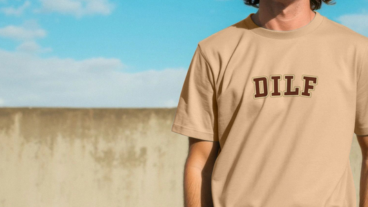 A person wearing a tan funny Dad T-shirt with "DILF" printed in bold letters stands outdoors against a backdrop of a blue sky and concrete wall.