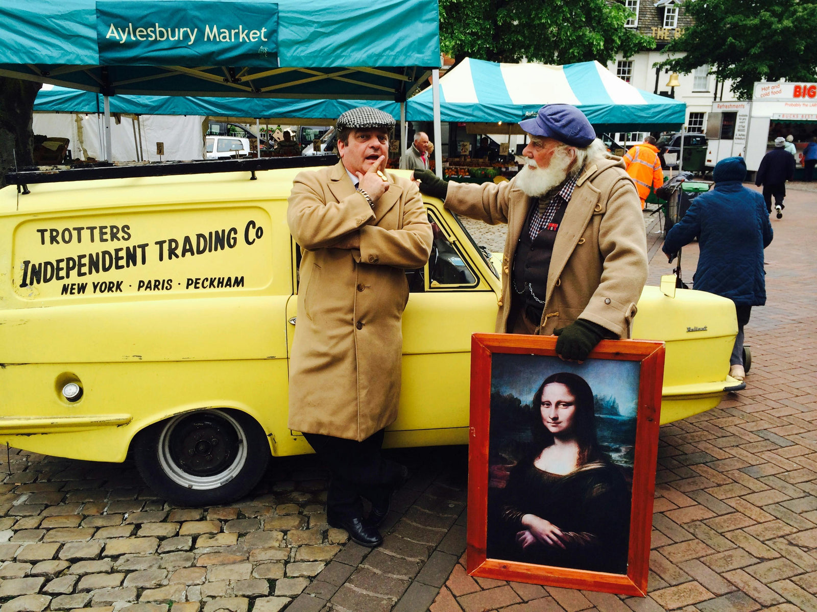 Two men stand outside a yellow van at Aylesbury Market. One wears a flat cap and trench coat, the other a cap and brown coat, holding a Mona Lisa print. The mood is playful.