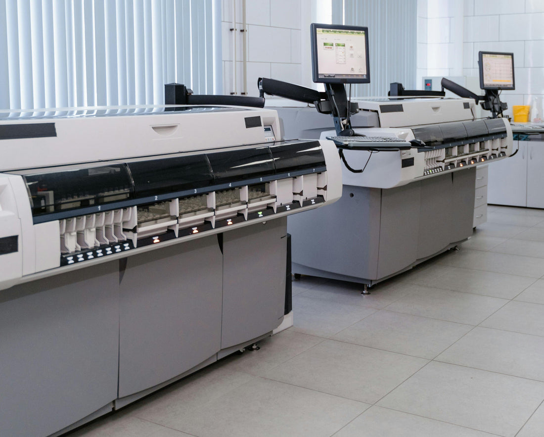 Two large industrial printers with control screens and keyboards in a bright, modern facility. The room has tiled floors and vertical blinds.