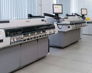 Two large industrial printers with control screens and keyboards in a bright, modern facility. The room has tiled floors and vertical blinds.