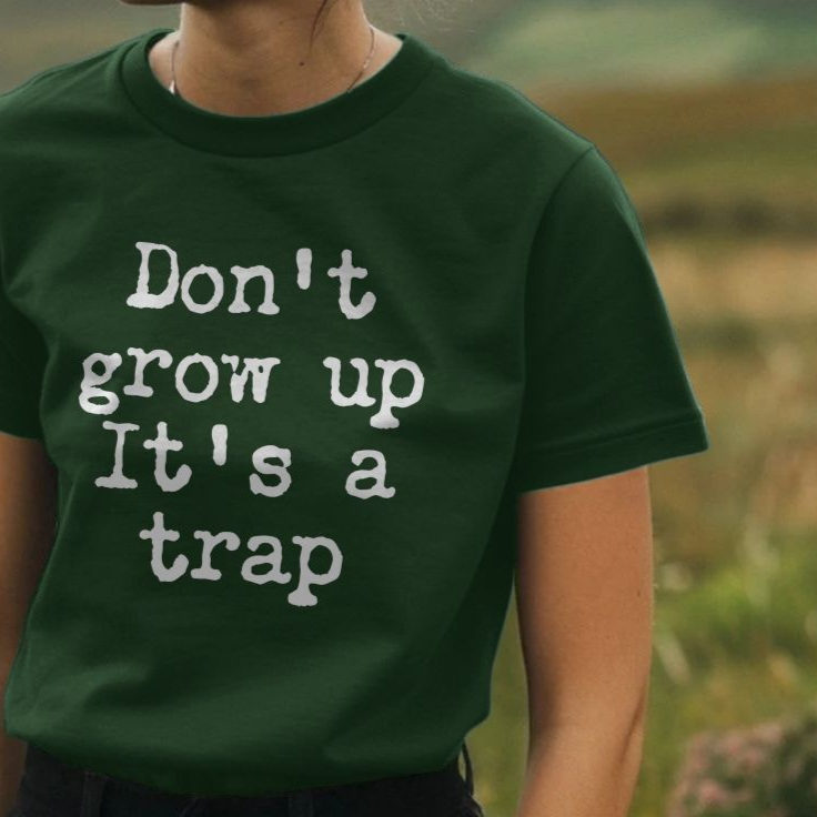 A person wearing a dark green Funny Slogan T-shirt with white text reading "Don't grow up, It's a trap" stands in a grassy field, conveying a playful tone.