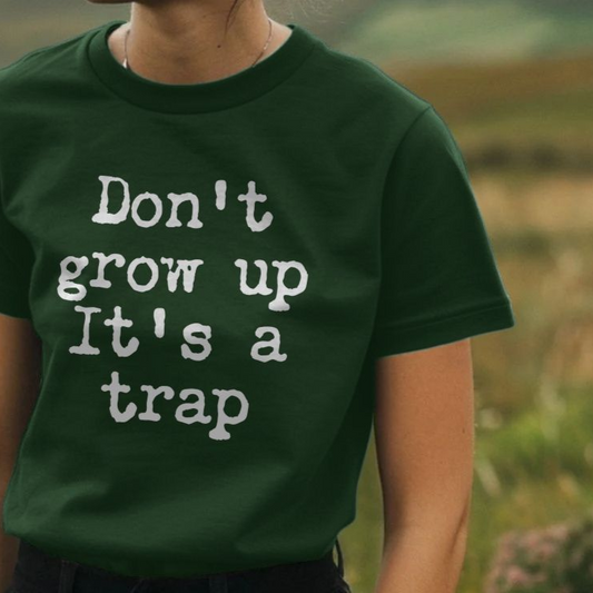 A person wearing a dark green Funny Slogan T-shirt with white text reading "Don't grow up, It's a trap" stands in a grassy field, conveying a playful tone.