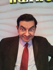 Wax figure of Mr Bean with exaggerated wide eyes and raised eyebrows. He's wearing a brown suit jacket, checkered shirt, and red tie, with a playful expression.