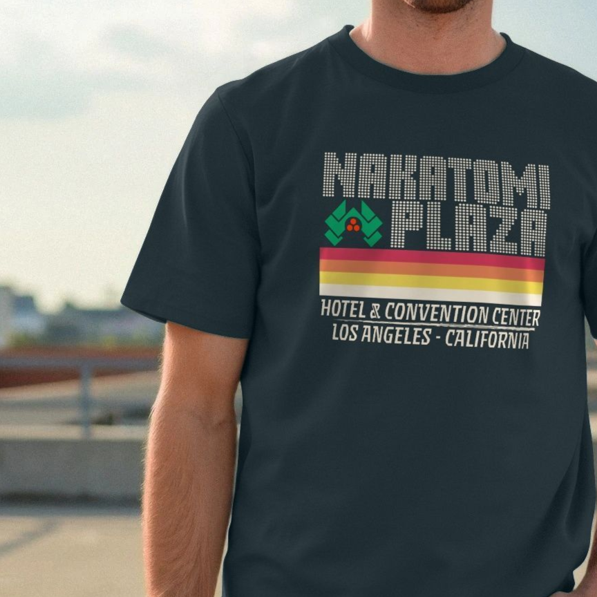 Man wearing a dark Christmas t-shirt with "Nakatomi Plaza" text, colorful stripes, and an abstract logo. The mood is casual and relaxed, outdoors setting.