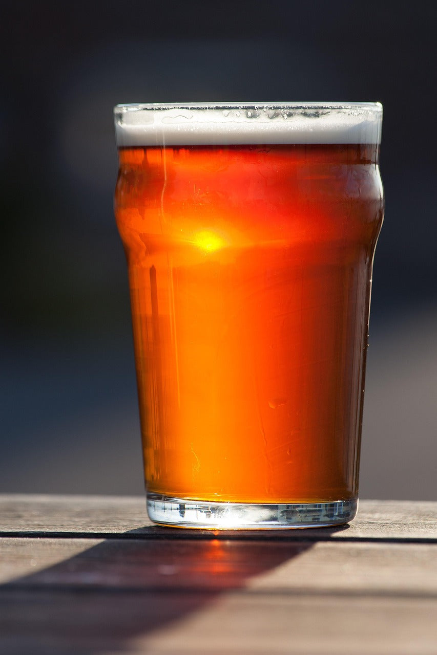 A pint of amber beer sits on a wooden table, sunlight illuminating the rich colour, with a frothy white head. Warm and inviting outdoor setting.