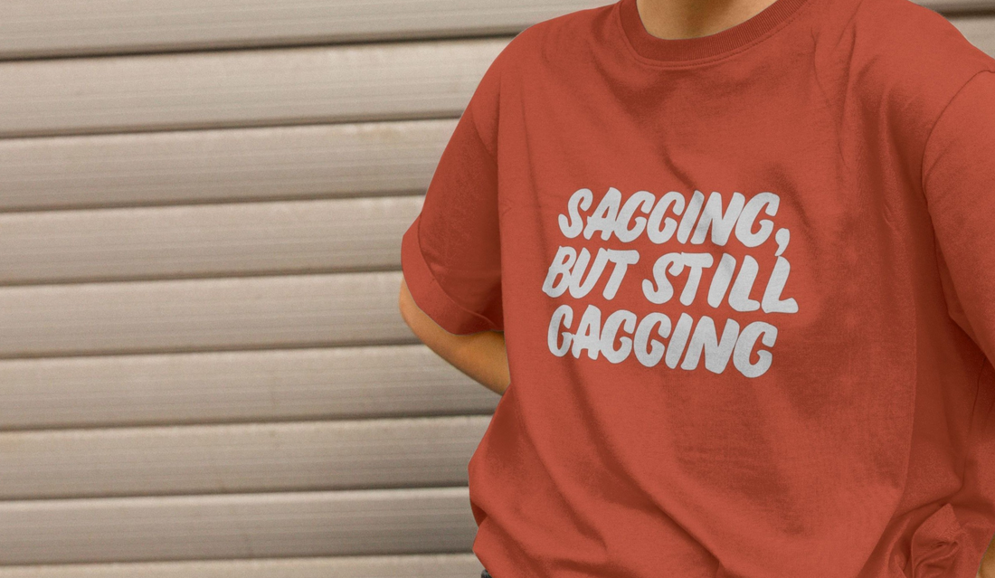 A person wearing a red offensive T-shirt with the playful phrase "SAGGING, BUT STILL GAGGING" in bold white letters. Background features a gray, horizontal metal shutter.