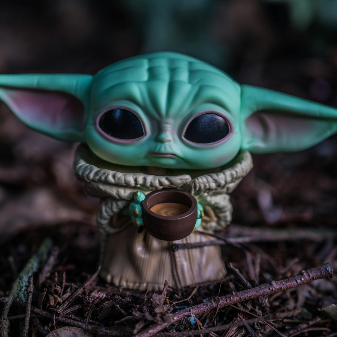 Toy figure of a small green creature with large ears holding a cup, set against a dark, forest-like background. It conveys a sense of calm curiosity.