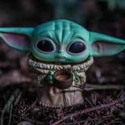 Toy figure of a small green creature with large ears holding a cup, set against a dark, forest-like background. It conveys a sense of calm curiosity.