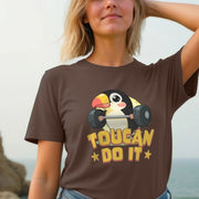 Smiling woman at a beach, wearing a brown T-shirt with a cute cartoon toucan lifting weights, captioned 