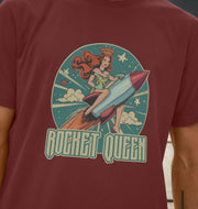 A person wears a dark red T-shirt featuring a retro-style illustration of a queen riding a rocket, with stars and clouds in the background. Text reads 