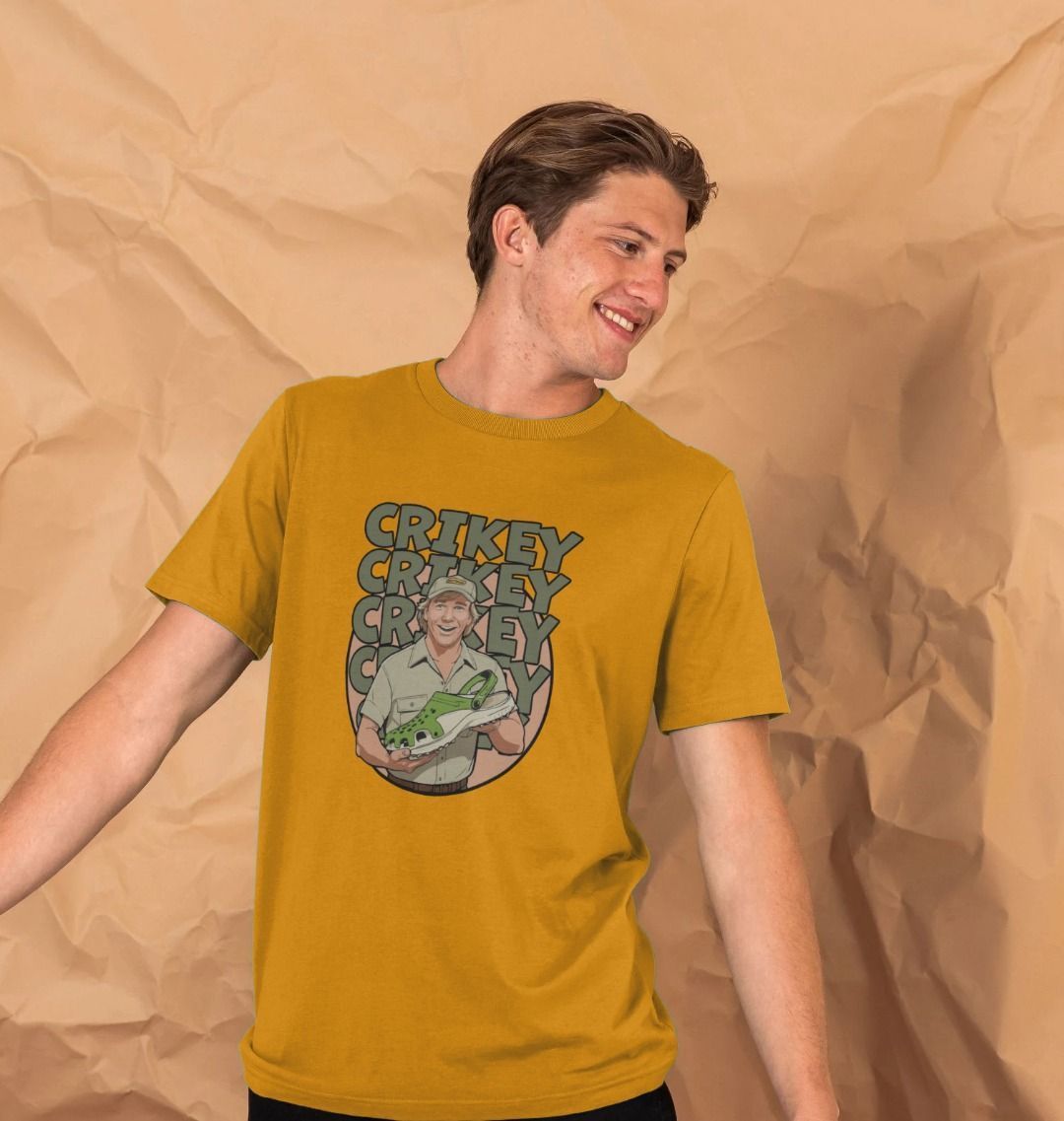 A smiling man in a mustard t-shirt stands against a beige, crumpled paper background. The shirt features Steve Irwin with the text "CRIKEY" repeated.