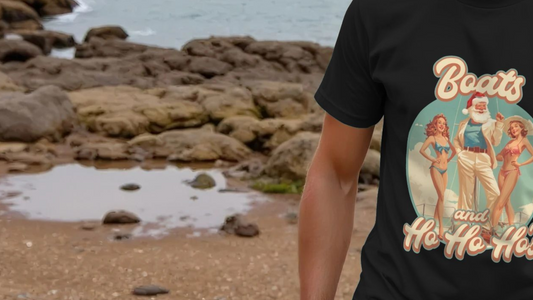 A person in a black Graphic Christmas T-shirt with a retro Step Brothers themed design of Santa and pin-up models stands by a rocky shoreline. The tone is playful and vintage.