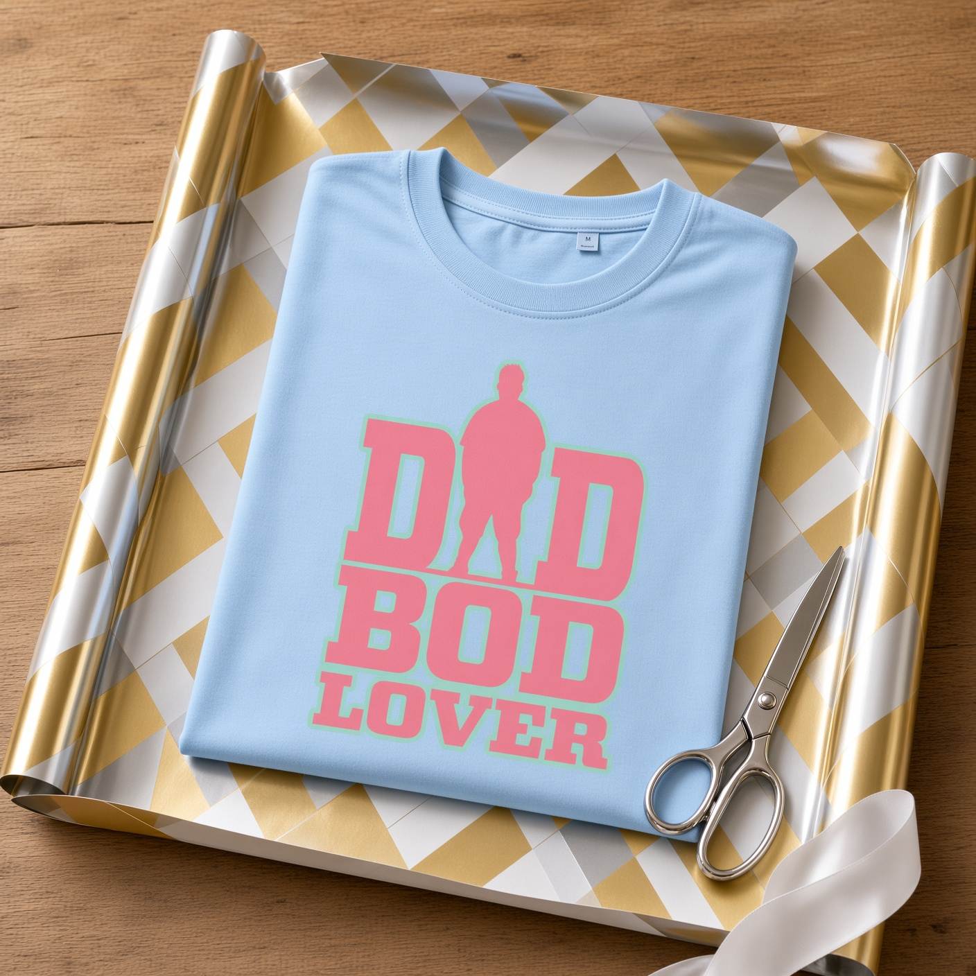 Sky blue "Dad Bod Lover" graphic t-shirt laid flat on top of gold and white checkered wrapping paper next to a pair of silver scissors