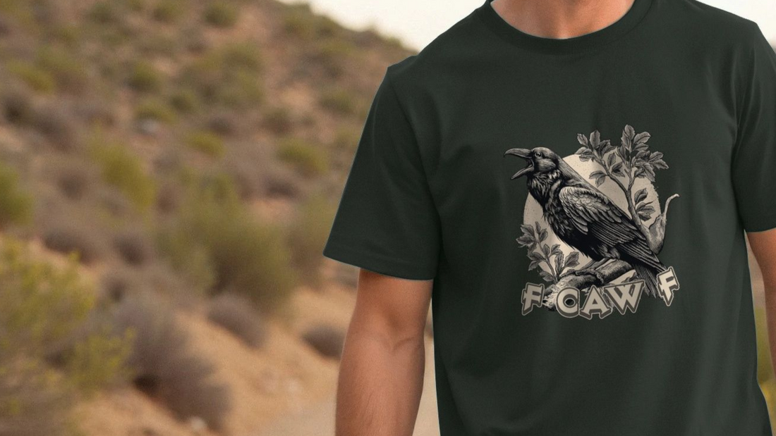A person wears a dark green funny graphic t-shirt with a detailed crow illustration perched on branches, with the witty slogan "Fcawf" against a blurry desert background. The tone is rugged and adventurous.