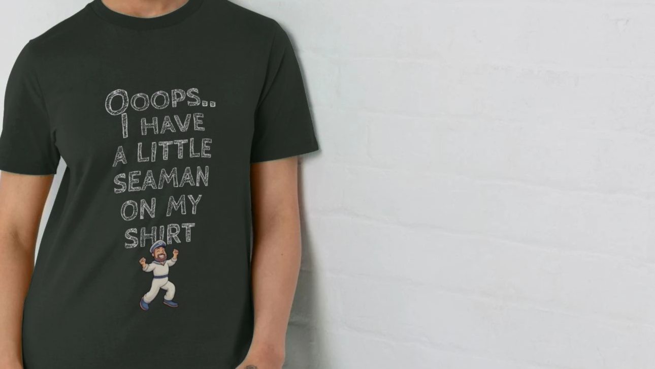 Woman wearing a black rude T-shirt with playful text "Ooops... I have a little seaman on my shirt" and a cartoon sailor illustration, set against a light wall.