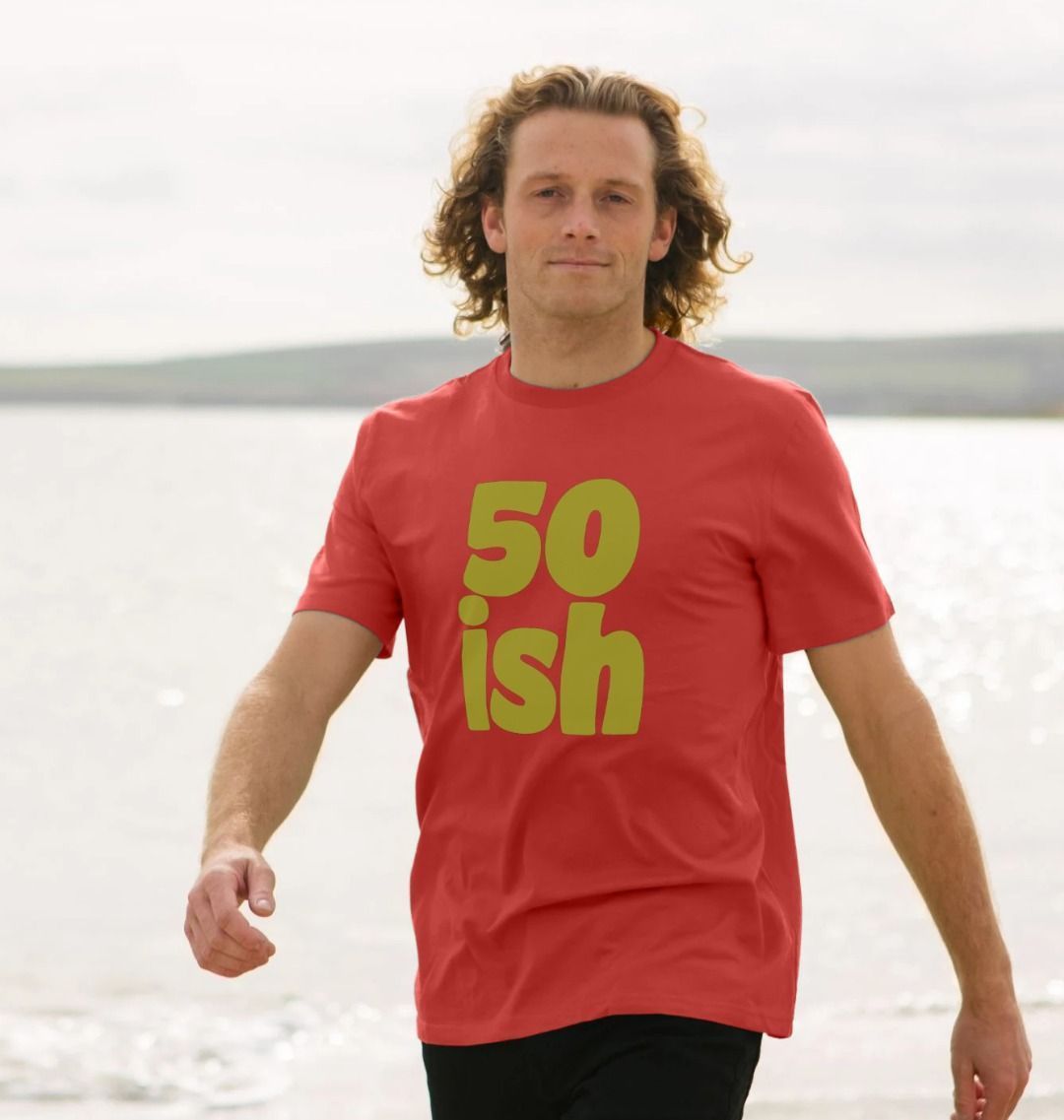 A man with curly hair walks on a beach, wearing a red shirt with "50 ish" in bold yellow. The background is a calm sea under a cloudy sky.