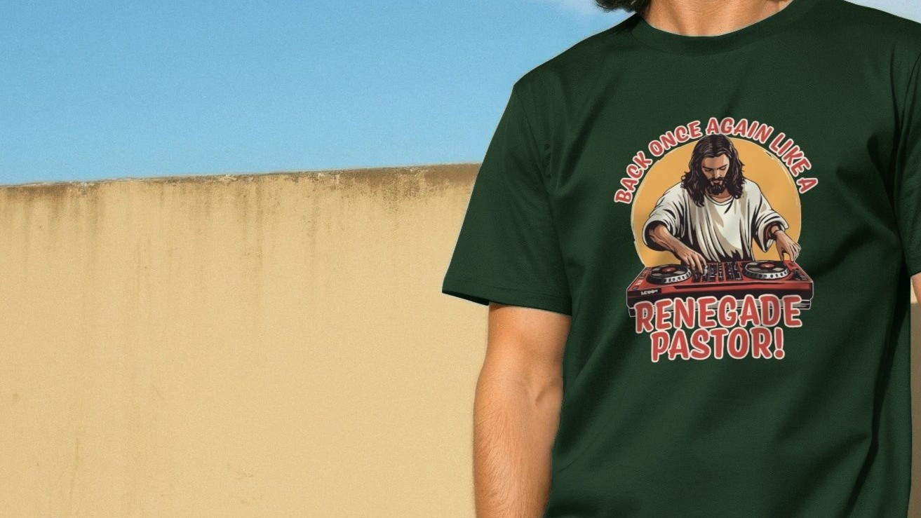 A person wearing a dark green funny T-shirt with a graphic of Jesus as a DJ and the words "Back once again like a renegade pastor!" against a beige wall.