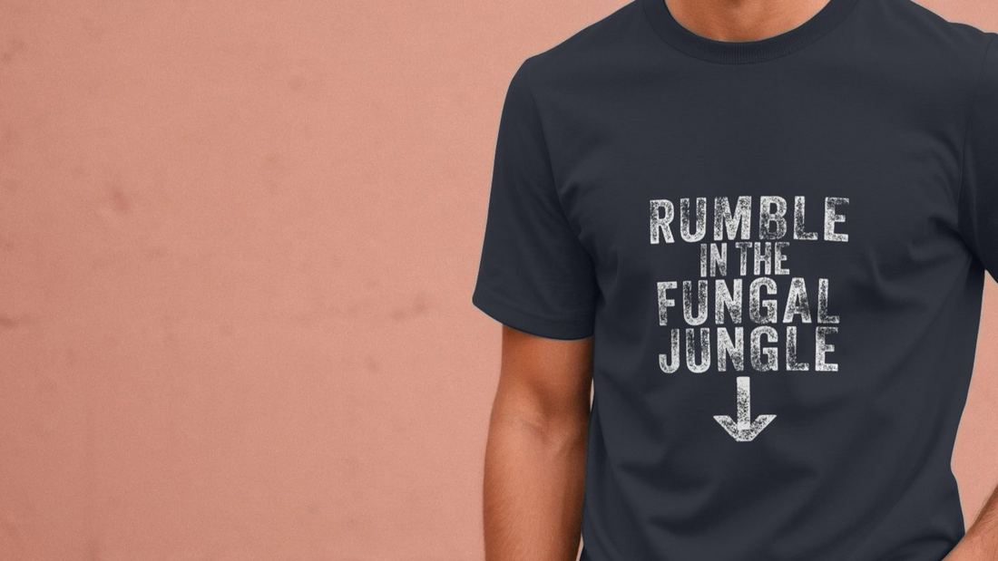 A person wearing a black funny slogan T-shirt with the white text "RUMBLE IN THE FUNGAL JUNGLE" and a downward arrow. The background is a muted pink wall.