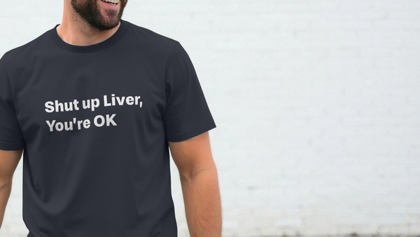 A man wearing a navy slogan T-shirt with the humorous text "Shut up Liver, You're OK" stands against a white brick wall, smiling. Tone is casual and playful.
