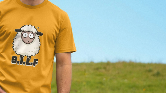 A person in a mustard-yellow funny animal T-shirt with a cartoon sheep and letters "S.I.L.F" stands against a grassy field. The sky is clear and blue, evoking a lighthearted, humorous tone.