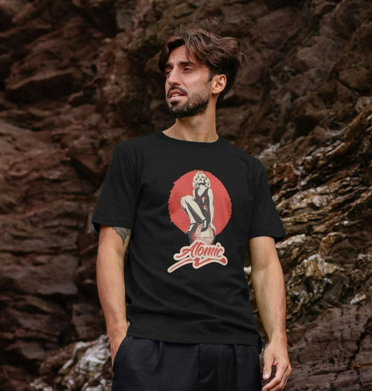 Man with short hair and a beard wearing a black "Atomic" T-shirt with vintage art, standing against a rocky background, looking to the side.