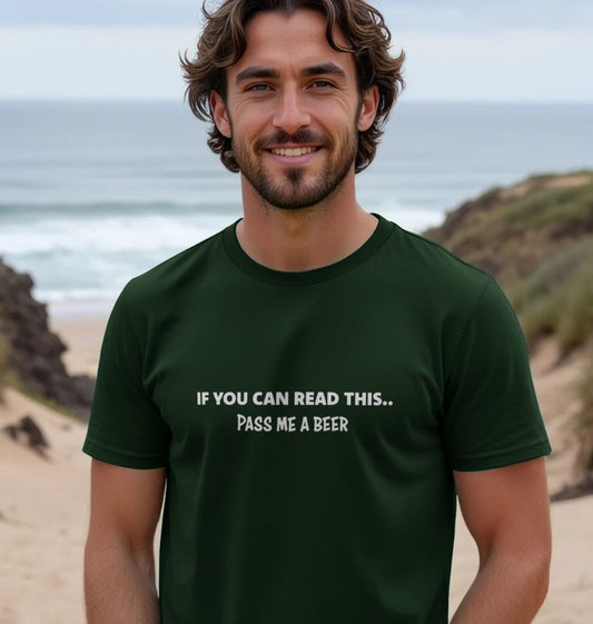 Smiling man on a beach wearing a dark green T-shirt with the joke text, "If you can read this... pass me a beer." The mood is light-hearted and casual.