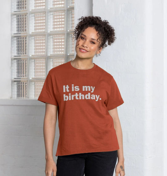 Woman smiles warmly, wearing a brick-red T-shirt with "It is my birthday." written on it. She's standing by a glass brick wall, exuding a cheerful vibe.