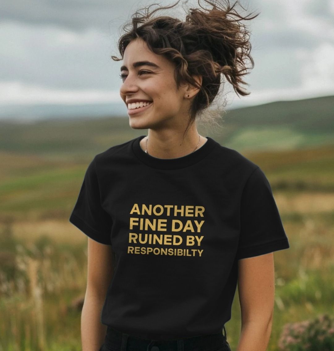 Young woman smiling in a windy field, wearing a black t-shirt with yellow text: "Another fine day ruined by responsibility." The tone is light and humorous.