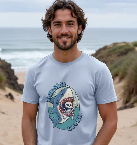 Smiling man on a beach wearing a light blue t-shirt with a cartoon shark design and humorous text. The ocean and sand dunes are in the background.