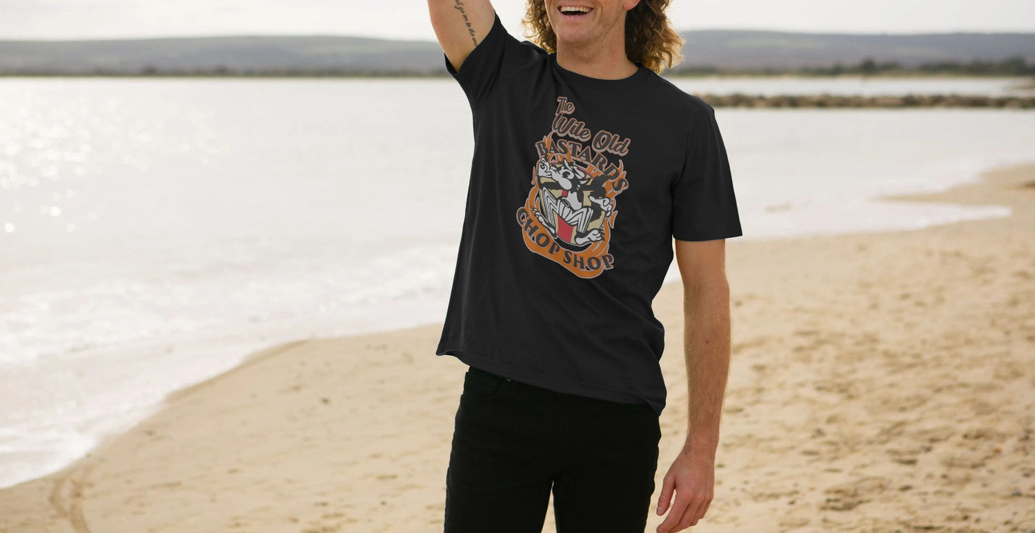 A man with long hair and a tattoo smiles while standing on a sunny beach. He wears a Motorcycle T-shirt with a cartoon design. The mood is relaxed and joyful.