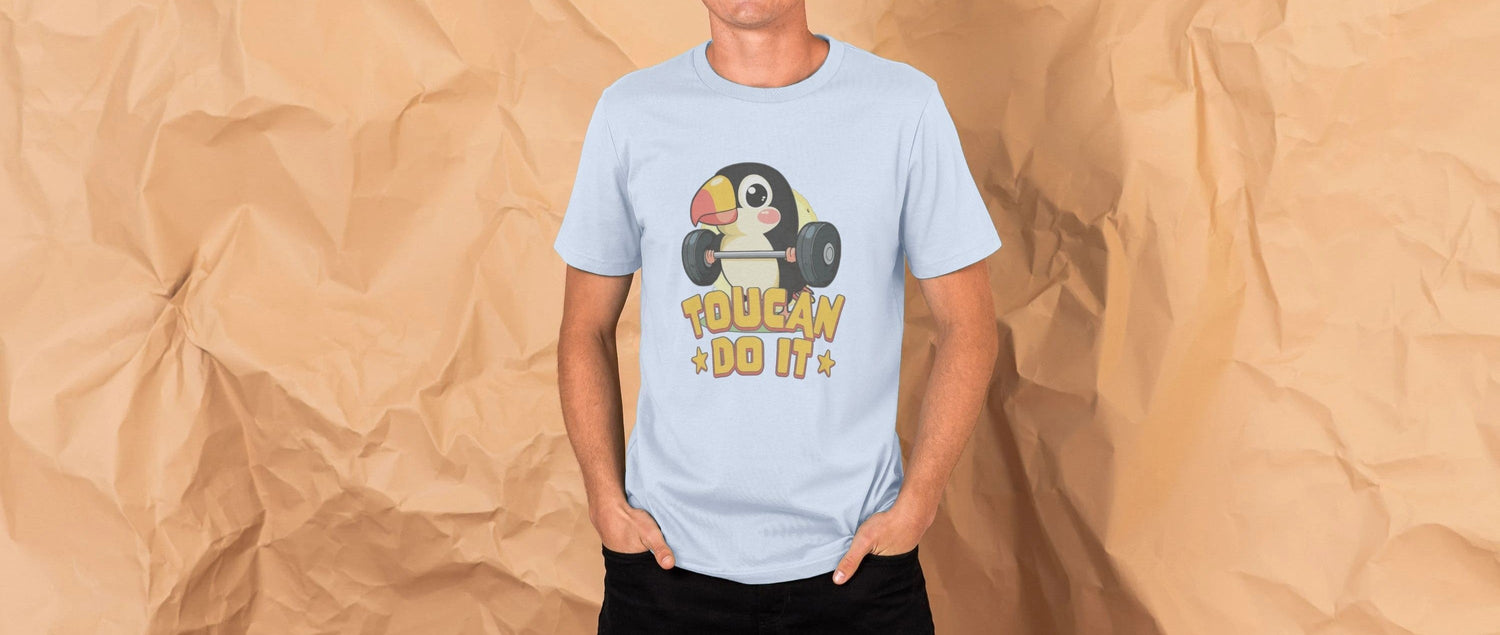 Man wearing a light blue Novelty T-shirt with a cartoon toucan lifting weights and the text "Toucans Do It." Background is crumpled brown paper.