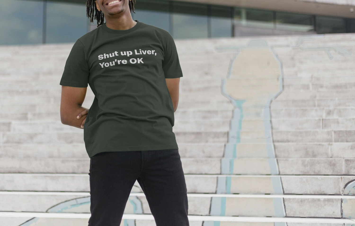 A smiling man stands on steps wearing a black Beer T-shirt with the text "Shut up Liver, You're OK." The scene conveys a playful, casual vibe.