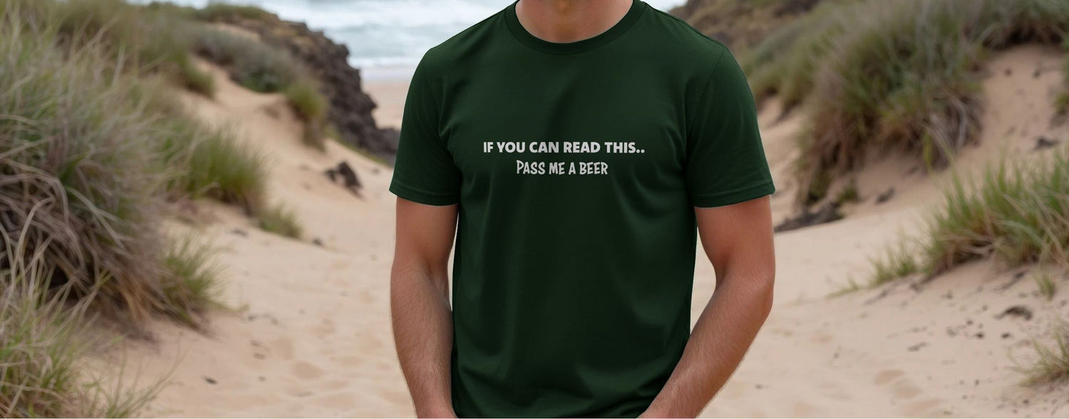 A person wearing a dark green beer T-shirt with the humorous phrase "If you can read this... pass me a beer" stands on a sandy beach path bordered by grass.